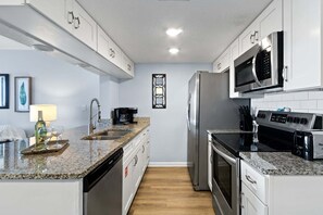 A modern kitchen with white cabinets, stainless steel appliances, granite countertops, and a wooden floor. - Beautiful 1BR Condo on Cape Canaveral Beach • Pool & Hot Tub 🌺 (Cape Canaveral)