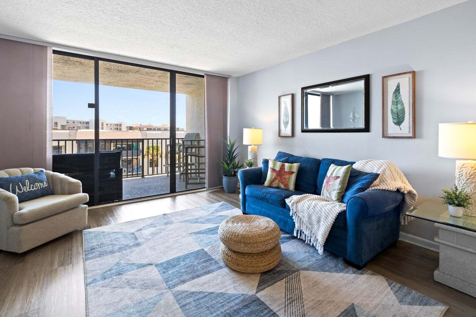 A living room with a sofa, a chair, a patterned rug, and a balcony with a view of buildings.