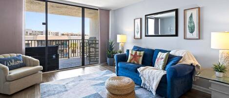 A living room with a sofa, a chair, a patterned rug, and a balcony with a view of buildings.