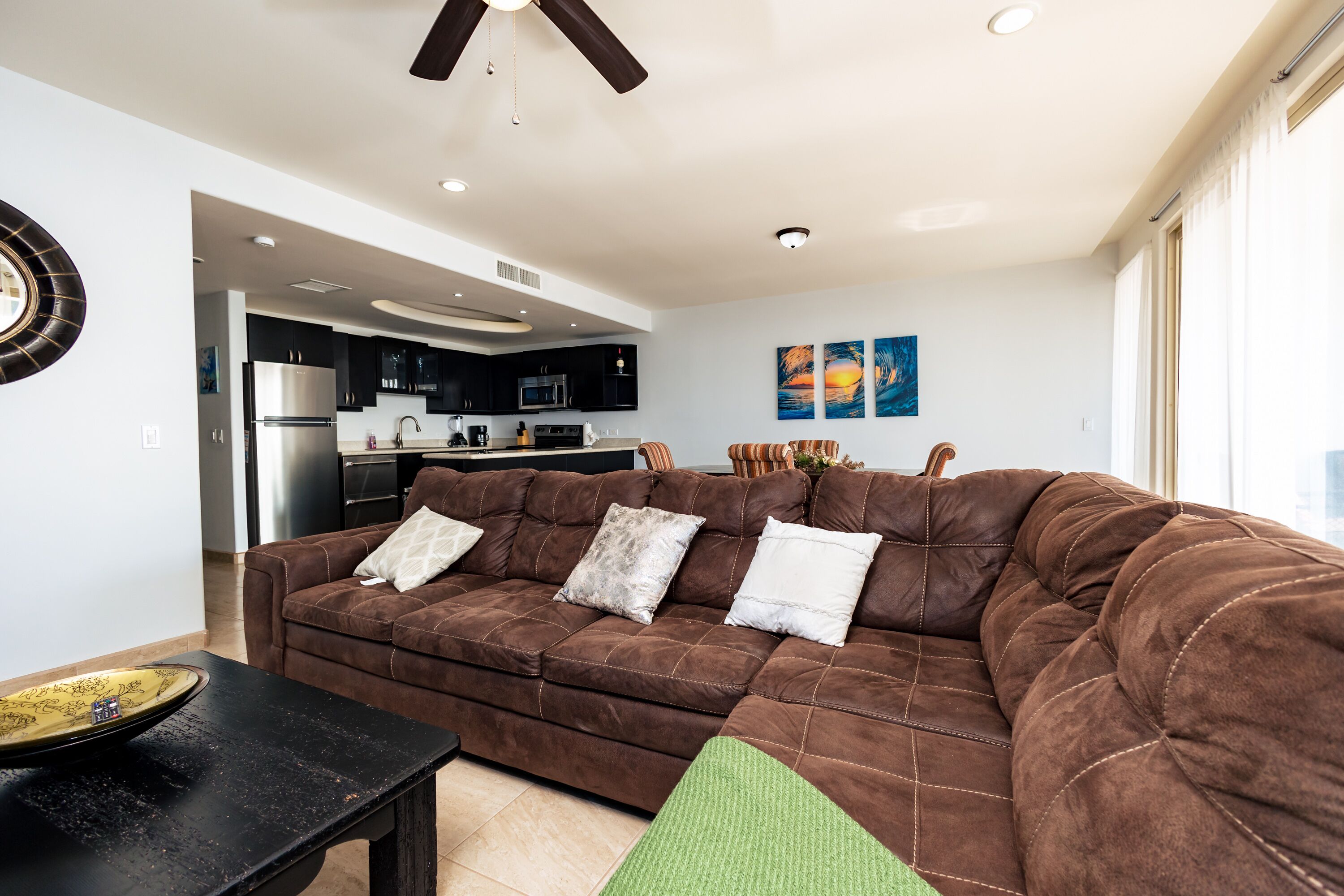 A living room with a brown sofa, a black coffee table, and a green chair. There is a kitchen in the background with dark cabinets and stainless steel appliances.