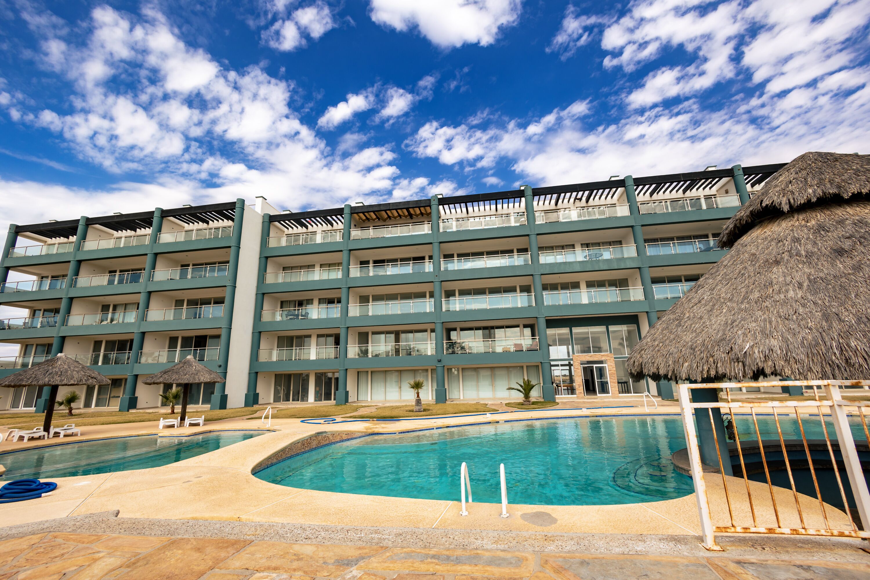 Et hotel med swimmingpool, flere balkoner og stråtag parasoller.
