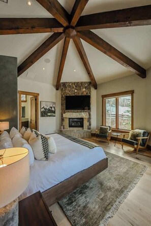 A bedroom with a large bed, a stone fireplace, a television, and a view of the outdoors.