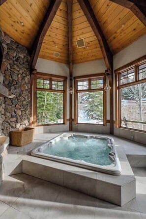 A hot tub in a room with stone walls and wooden beams, large windows, and a view of trees. - Abode at Northside, ski-in/ski-out, hot tub, mountain views (Park City)