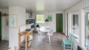 Eine kleine Küche mit weißen Schränken, ein Essbereich mit rundem Tisch und Stühlen sowie ein Blick ins Freie durch ein Fenster.