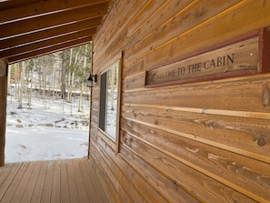 Eine Holzhütte mit einem Schild 'WELCOME TO THE CABIN', einer Holzterrasse und Blick auf einen verschneiten Wald.