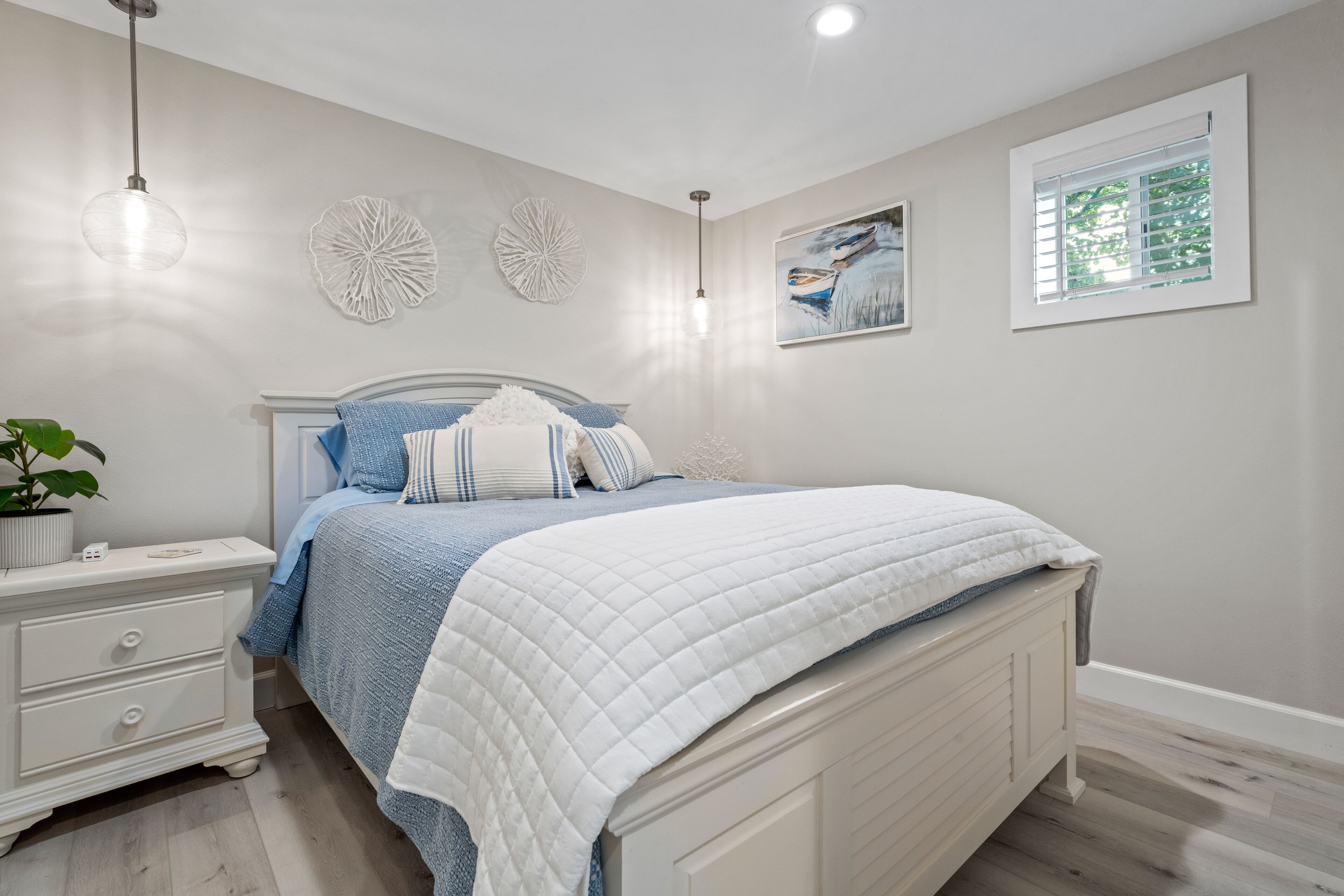 A bedroom with a bed, bedside tables, a nightstand, a potted plant, wall decor, and a window with blinds.