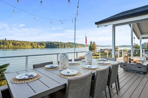 An outdoor dining area with tables set for two, overlooking a lake with a forested shoreline. - Grapeview- Luxury Waterfront Home w/ Hot Tub (Grapeview)