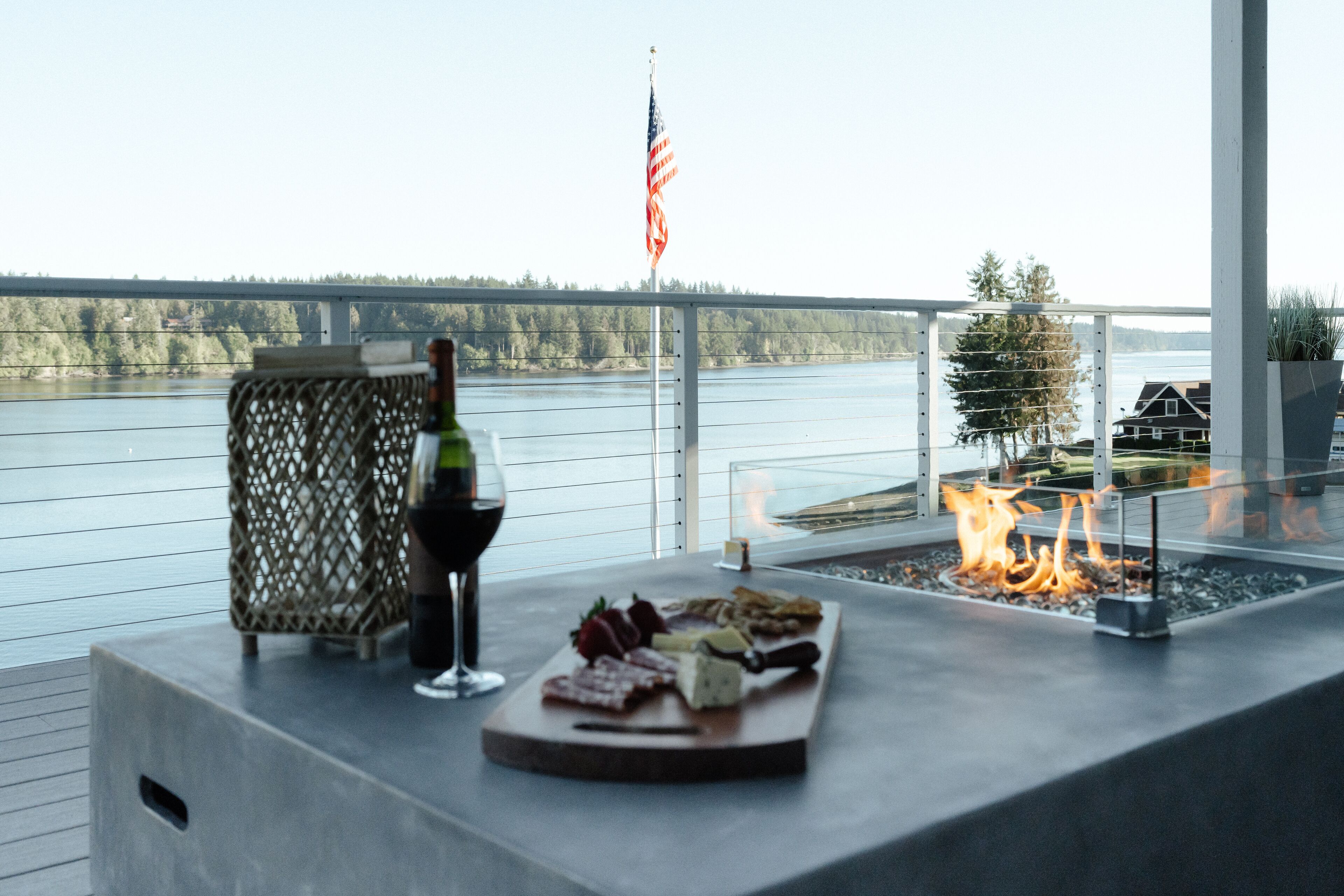 Une terrasse agrémentée d’un foyer, une bouteille de vin et une planche de charcuterie, le tout offrant une vue sur un plan d’eau avec un pont et des arbres en arrière-plan.
