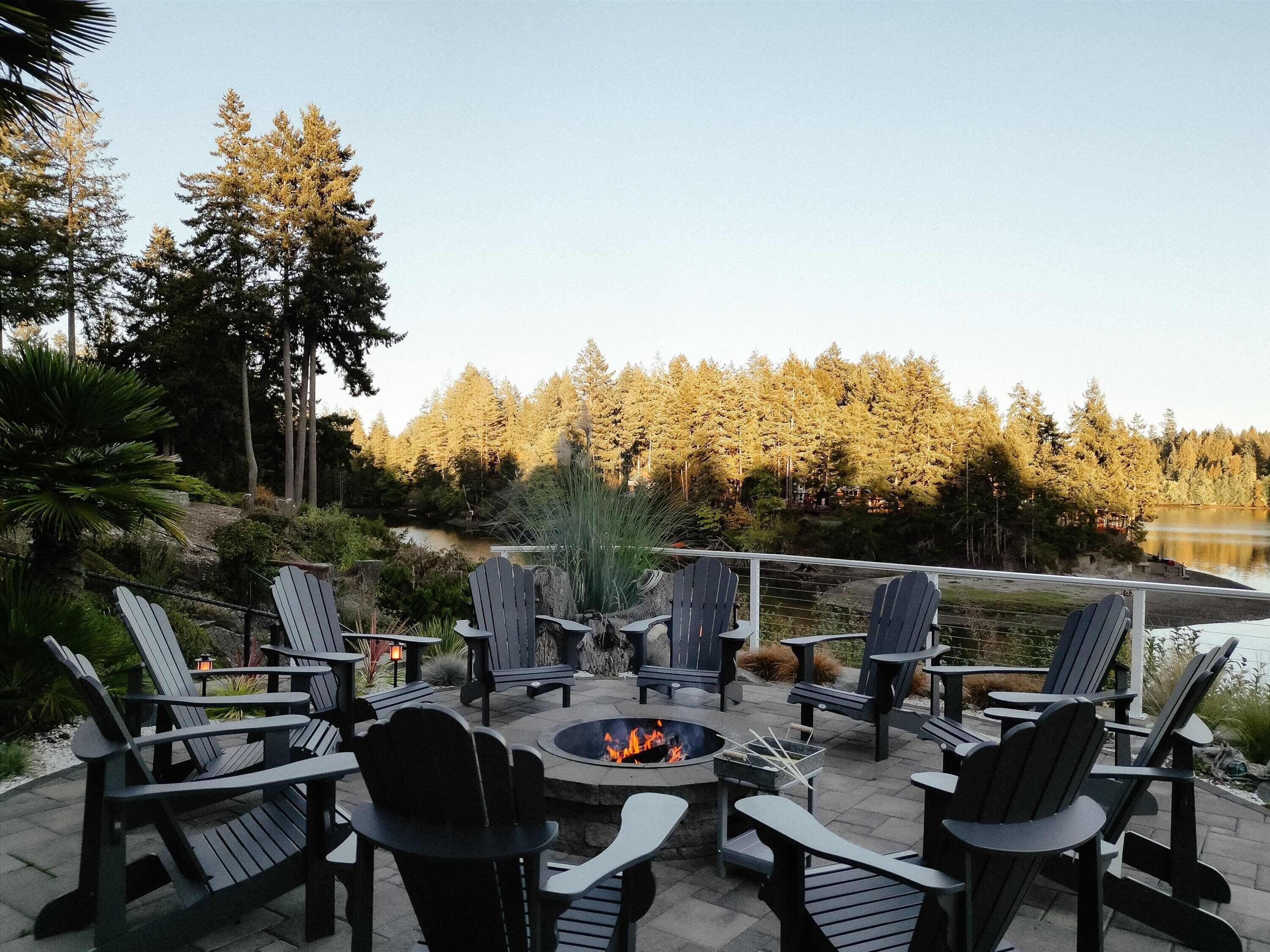 An outdoor seating area with Adirondack chairs and a fire pit, overlooking a lake and forested area.