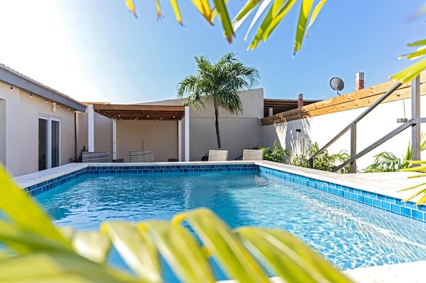 A swimming pool with a tiled edge, surrounded by a covered patio area with seating. - Ojanea Home- 3BR- Spacious w/Pool/Perfect location (Oranjestad-West)