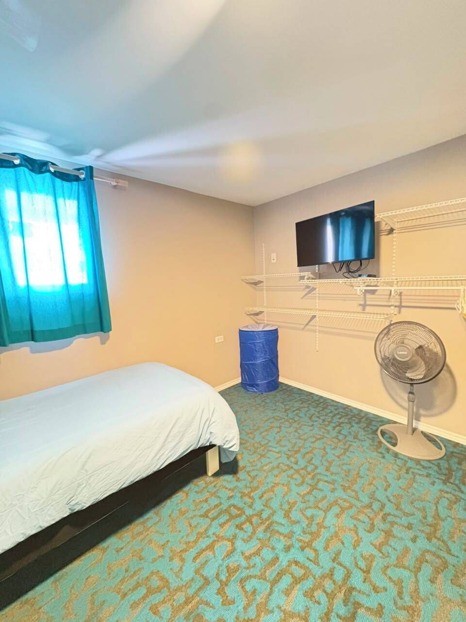 A bedroom with a bed, a TV mounted on the wall, a fan, and a blue barrel.