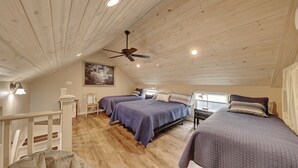 Una camera da letto mansardata accogliente con travi a vista in legno, due letti con biancheria viola, una ventola a soffitto e un quadro appeso al muro.