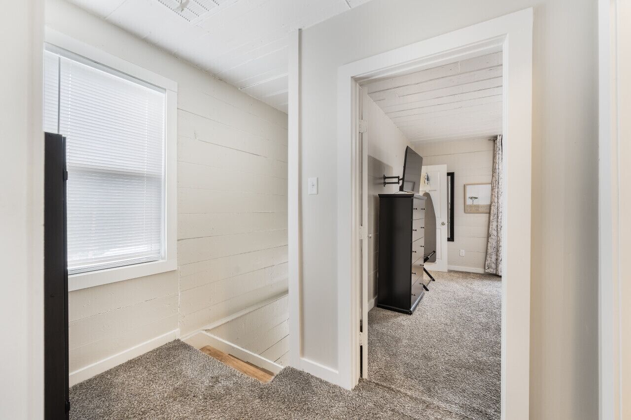 A hallway with white walls, a door leading to a room with a black cabinet and a mirror, and a window with blinds.