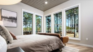 Een slaapkamer met een groot bed, uitzicht op bomen door grote ramen en een ingelijst schilderij aan de muur.