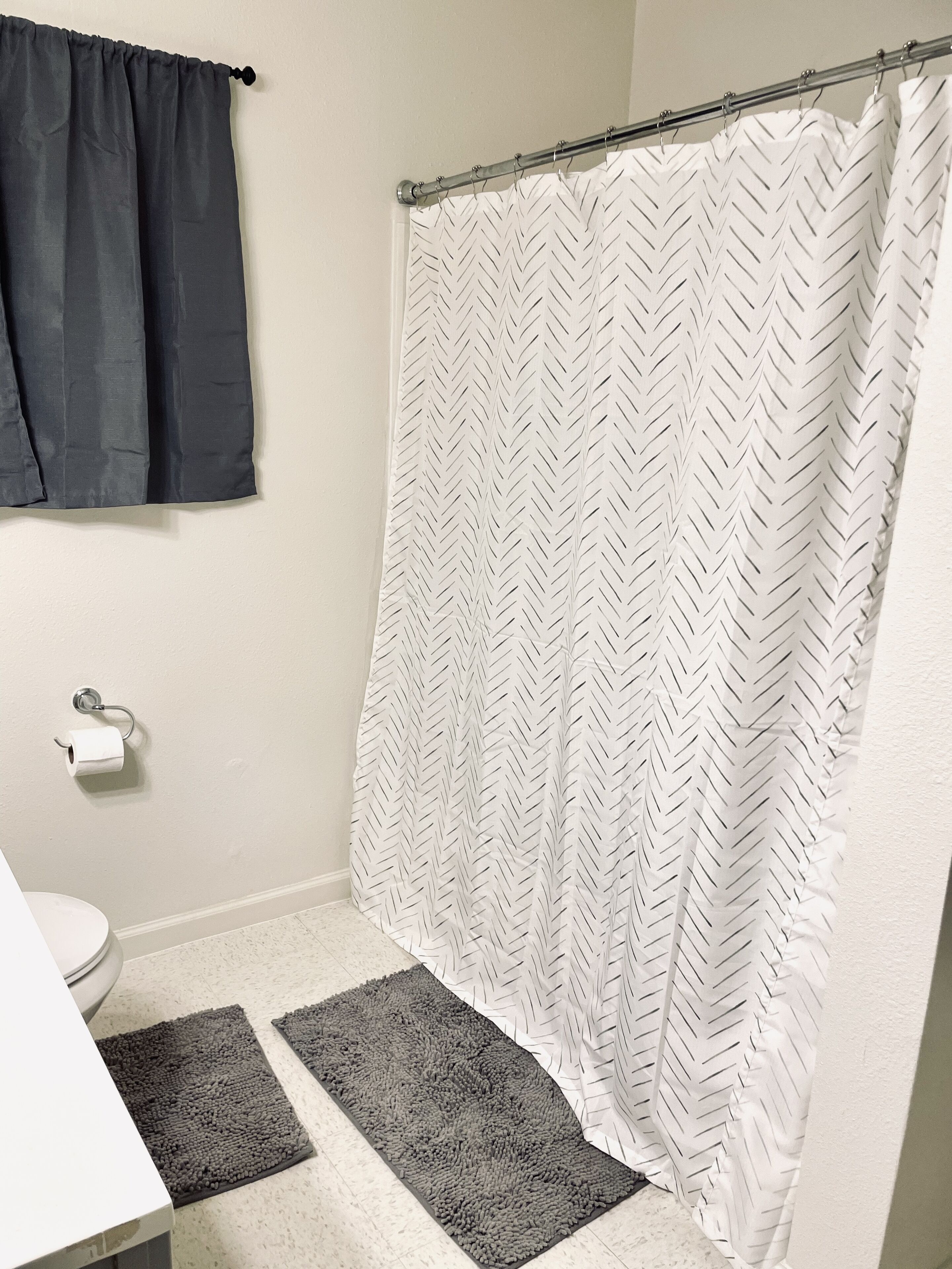 A bathroom with a shower curtain, toilet paper, and two rugs.
