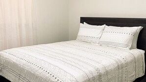 Un letto rifatto con cura, con copriletto e cuscini a fantasia, una testiera scura e una finestra con tende trasparenti.