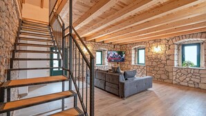 Un'ampia zona giorno con scala in legno, pareti in pietra e un divano grigio.