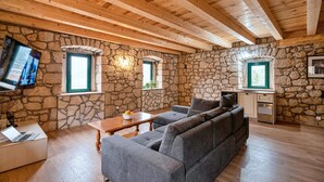 Una camera accogliente con pareti in pietra, soffitto in legno, un divano grigio, un tavolo in legno e un televisore fissato alla parete.