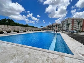 A swimming pool with lounge chairs, a stone wall, and a clear blue sky. - Brand New Punta Cana 2 Bedroom 2 B Condo (Punta Cana)
