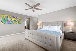 A bedroom with a tufted headboard bed, a ceiling fan, a nightstand with a lamp, and a painting on the wall.