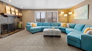 A living room with a sectional sofa, a fireplace, and a large window with blinds.