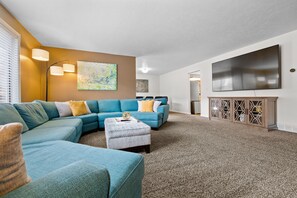 A living room with a sectional sofa, ottoman, and a flat-screen TV mounted on the wall.