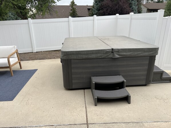 A hot tub with a built-in seat and a step for easy access, placed on a concrete patio with a white fence in the background.