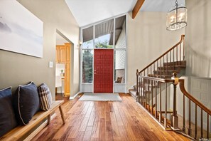 Una entrada con piso de madera, una puerta roja, un banco con cojines y una escalera con barandilla de madera.
