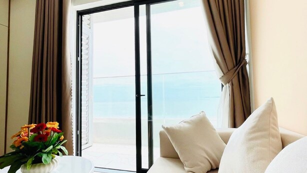 Ein modernes Zimmer mit einer Couch, einem kleinen Tisch mit Blumenvase und einer großen Schiebeglastür, die auf einen Balkon mit Meerblick führt.
