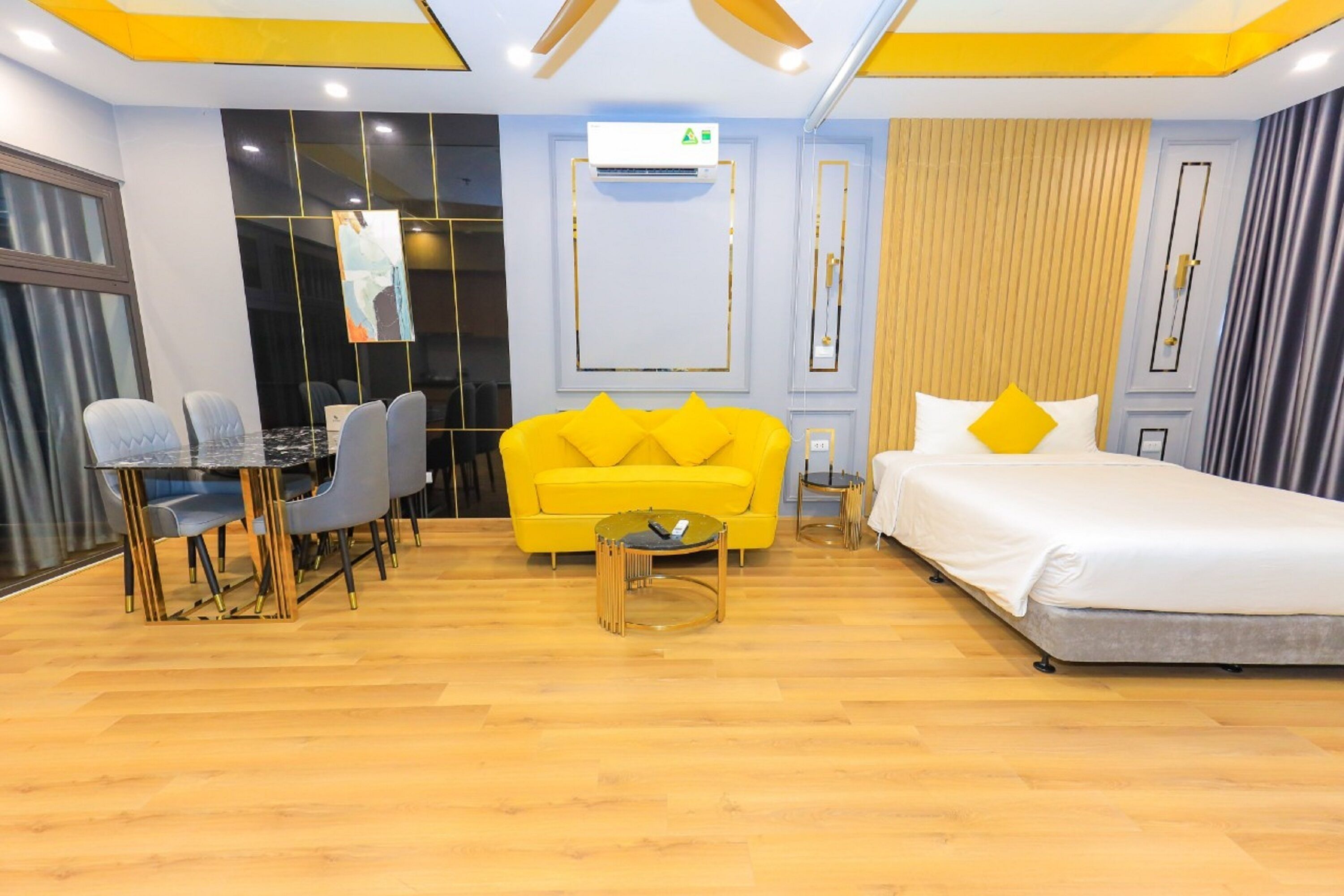 Quarto de hotel com piso de madeira, sofá amarelo com almofadas amarelas, cama branca, mesa redonda pequena e escrivaninha com cadeiras.