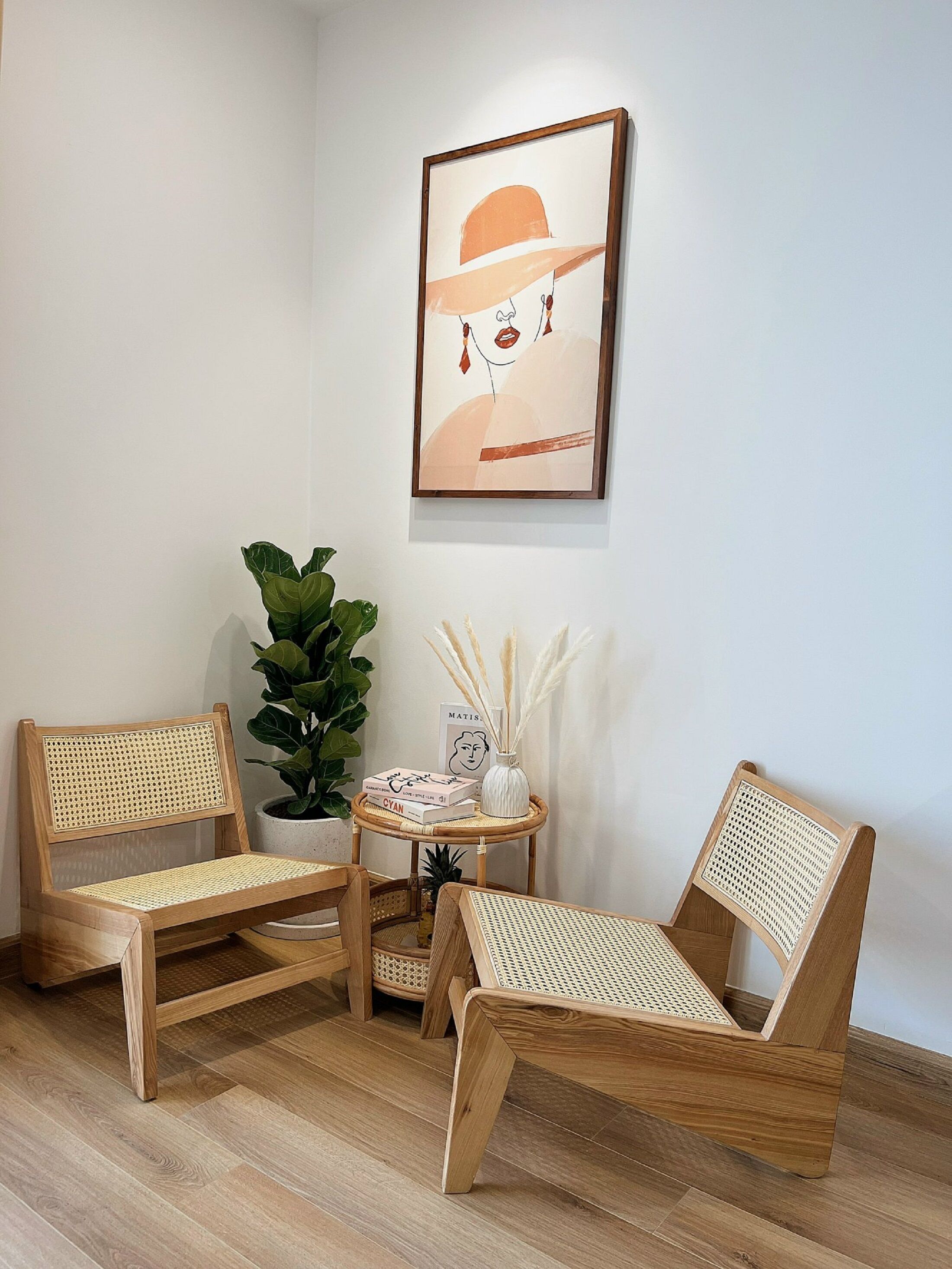 Ein Zimmer mit zwei Korbstühlen, einem kleinen runden Tisch mit Büchern und einer Vase, einer Zimmerpflanze und einem gerahmten Kunstwerk an der Wand.