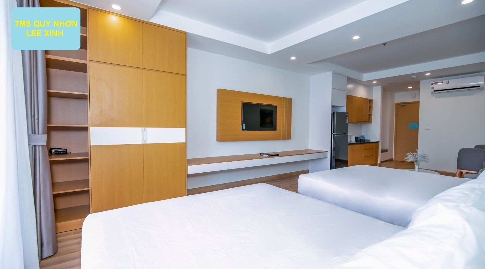 A hotel room with two beds, a wooden wardrobe, a small kitchenette, and a mounted TV.