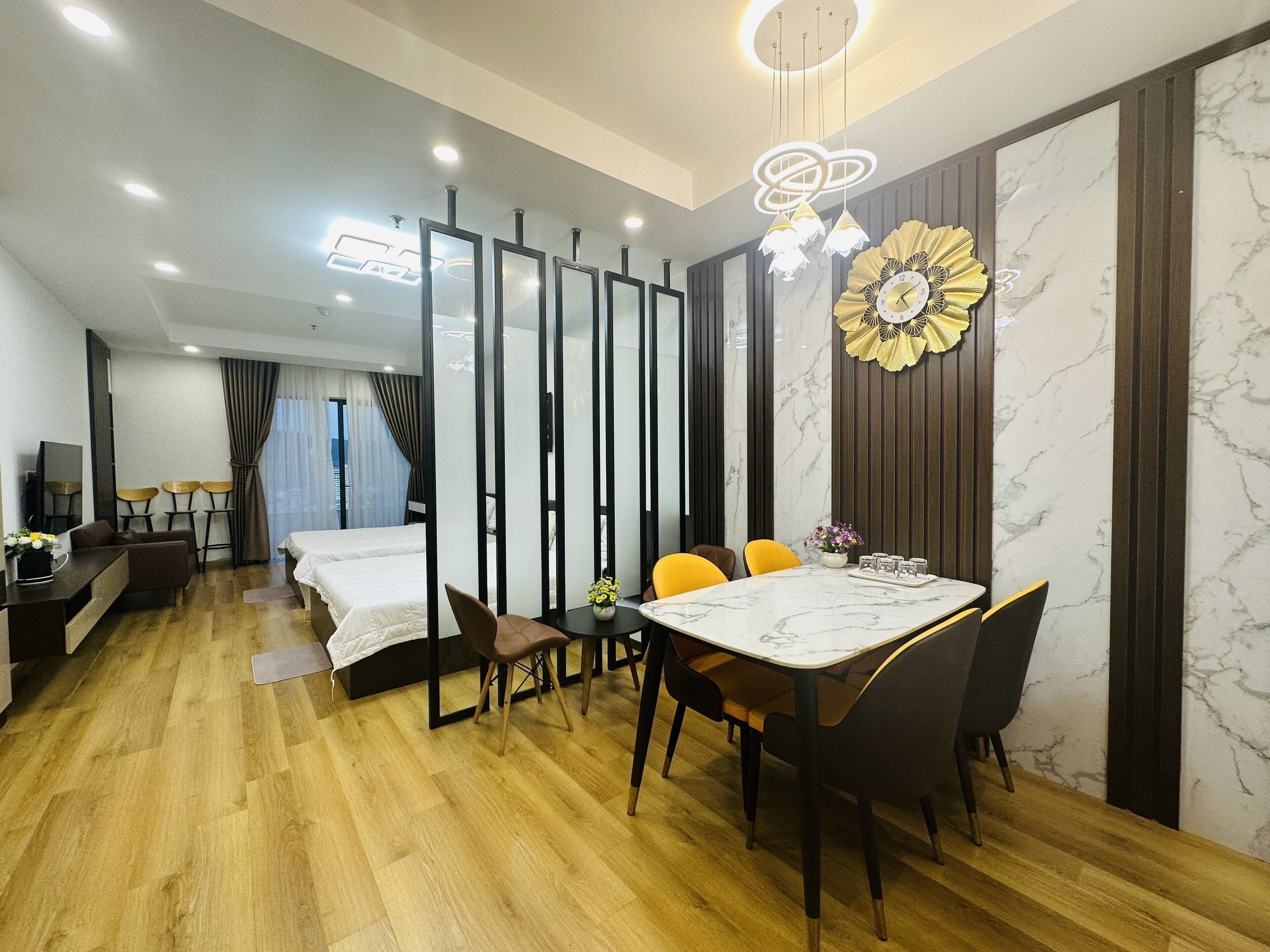 A hotel room with a wooden floor, a dining area with a marble table, two beds, a TV, and a decorative wall piece.
