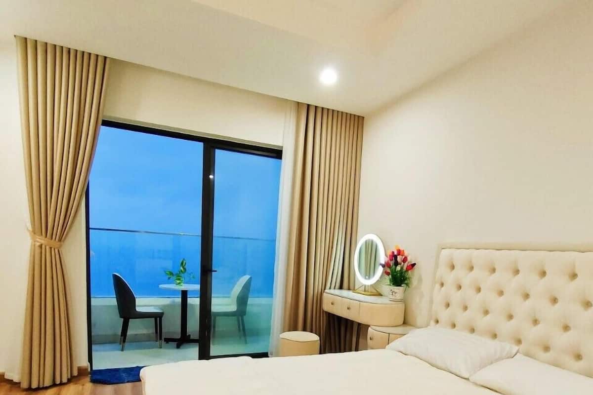 A hotel room with a large bed, a bedside table with a mirror, a small table with chairs, and a view of the outside.