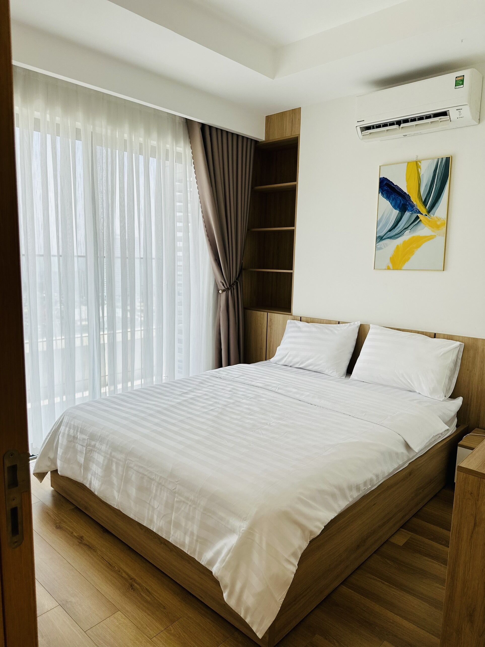 A modern bedroom with a wooden bed frame, white bedding, a large window with sheer curtains, a wall-mounted air conditioner, and a colorful abstract painting.