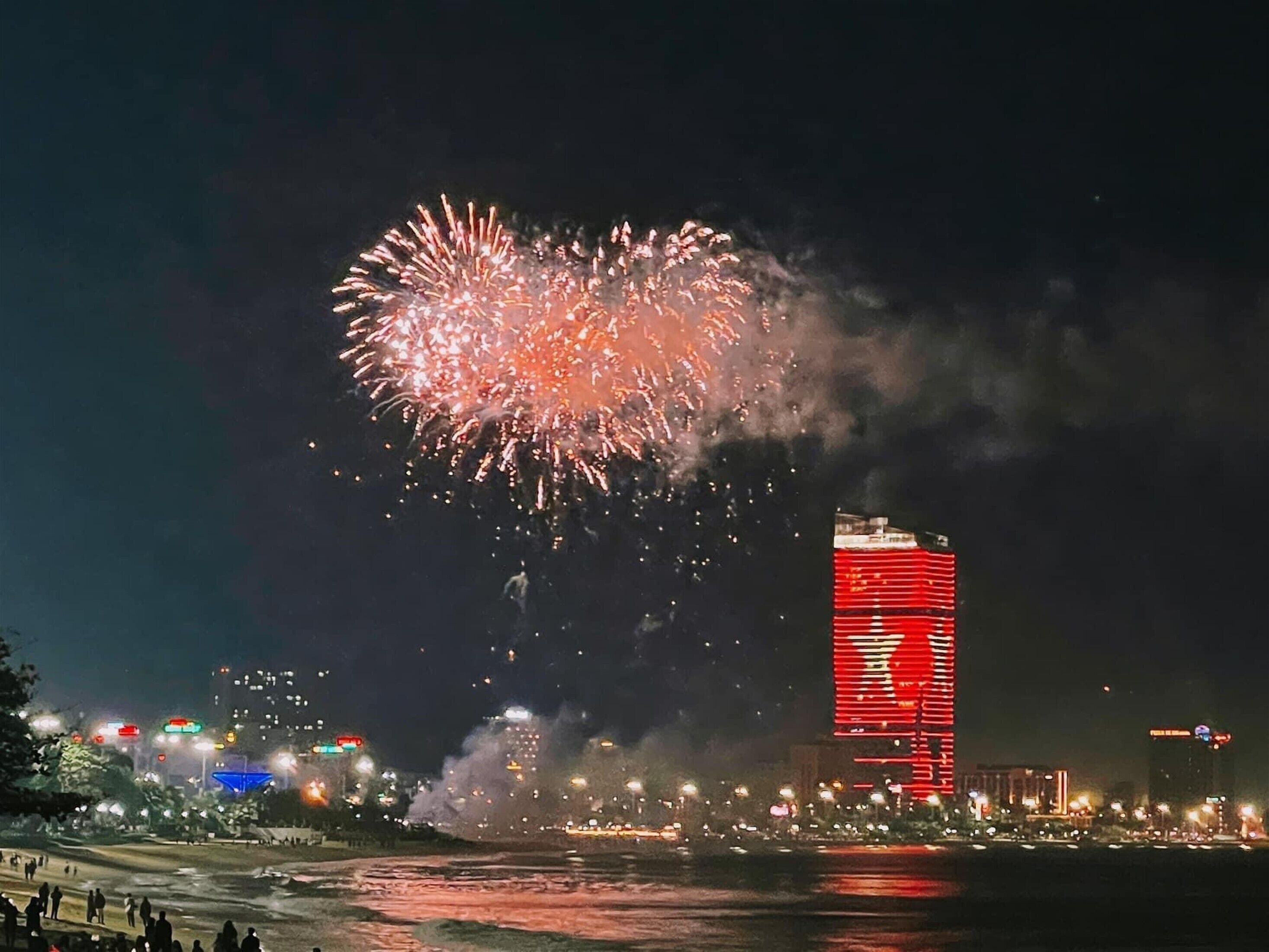 Une vue nocturne d’une ville, illuminée par des feux d’artifice, avec un bâtiment imposant aux lumières rouges et une zone riveraine.