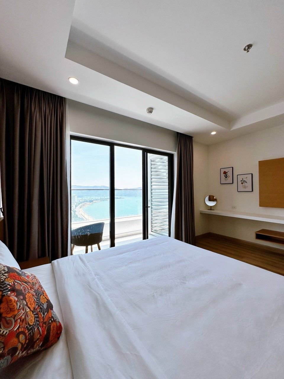 A hotel room with a large bed, a view of the sea, and a balcony with a chair.