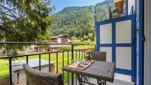 A balcony with wicker furniture, a table with a tray, and a view of a mountain and buildings. - V Balme 02: Beside Ski Lift; Pool, Sauna & Gym, Vallorcine (Argentiere), France (Vallorcine (Argentiere))