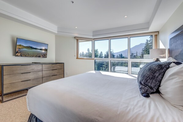 A bedroom with a large bed, a dresser, a TV on the wall, and a view of a mountain through large windows. - Le Chamois 504 - Upper Village (Whistler)