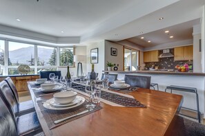 A modern dining area with a wooden table set for a meal, large windows offering a mountain view, and a kitchen with wooden cabinets and a tiled backsplash.
