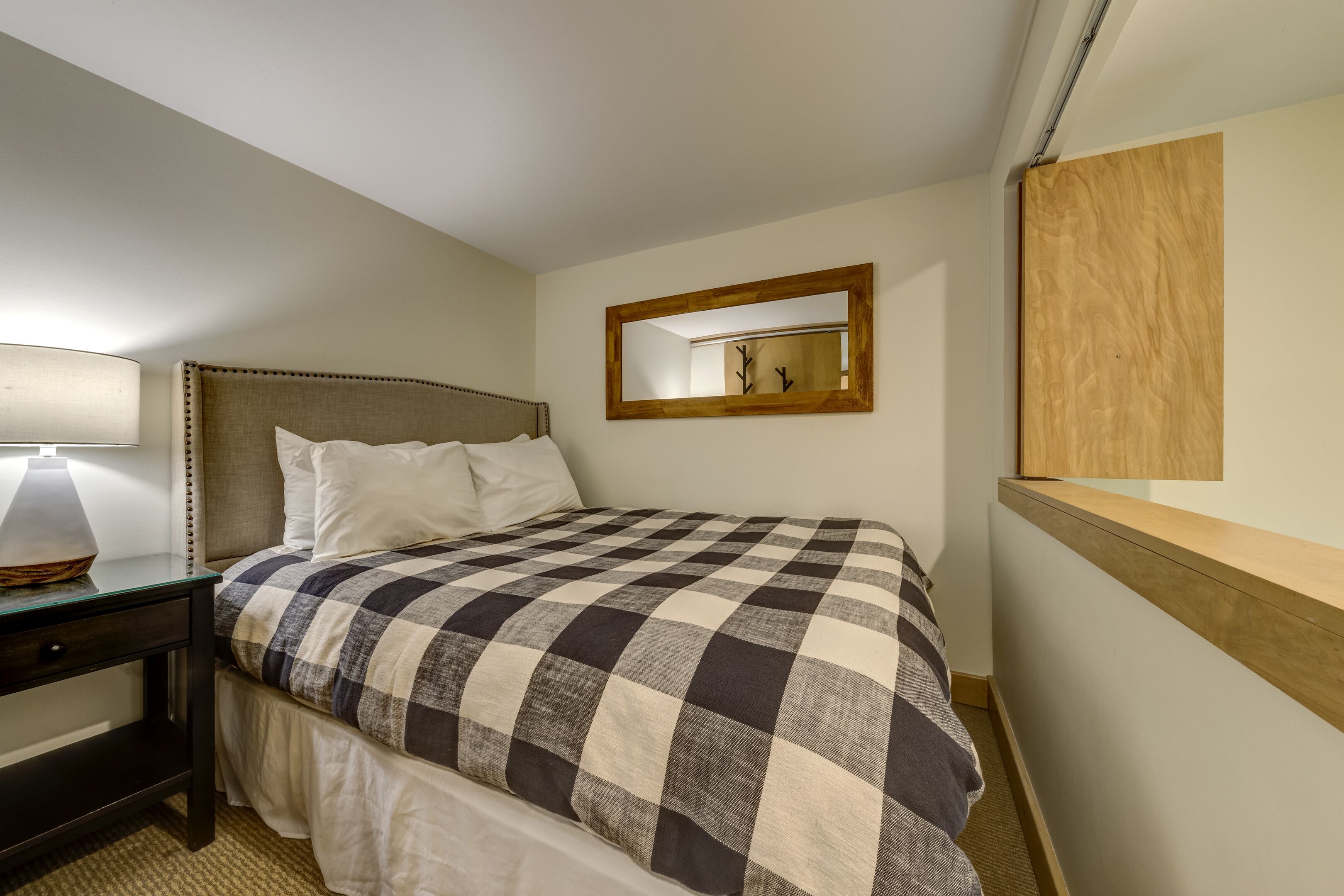 A neatly made bed with a checkered bedspread, a bedside table with a lamp, and a mirror on the wall.