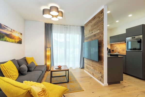 Una sala de estar moderna con un sofá gris, cojines amarillos, una mesa de centro y un televisor. El cuarto tiene piso de madera y un ventanal con cortinas.