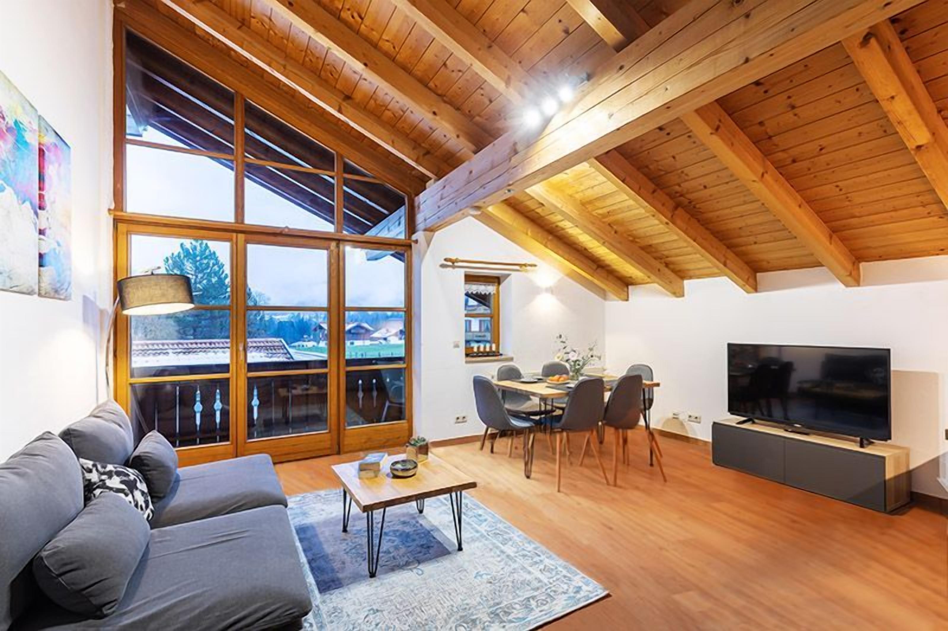 Een moderne woonkamer met een houten plafond, een bank, een salontafel, een televisie, en een eethoek met stoelen en een tafel.