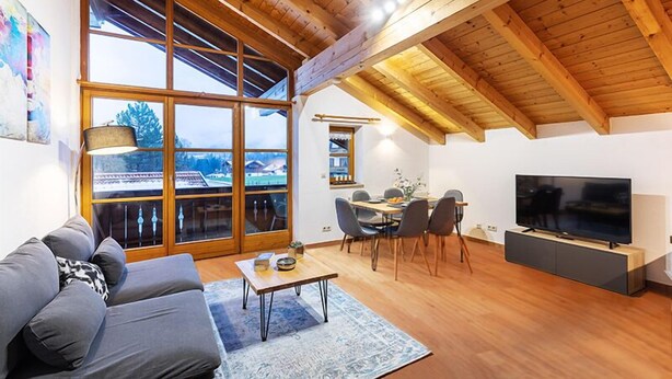 Uma sala de estar ampla com uma janela grande, vigas de madeira no teto, um sofá, uma mesa de jantar com cadeiras e uma TV de tela plana.