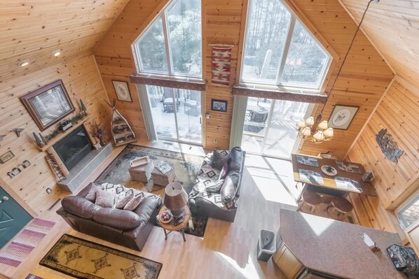 A cozy cabin interior with a fireplace, a sofa, a dining table, and large windows offering a view of the snowy outdoors.