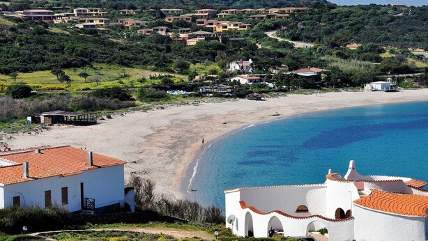A coastal area with a sandy beach, a clear blue sea, and a hillside with numerous houses.