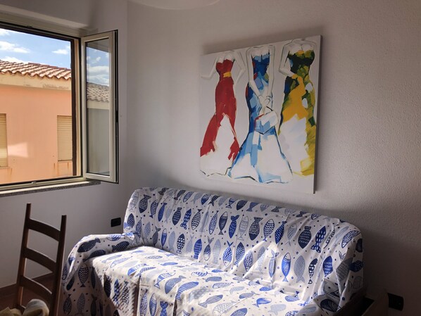 Une pièce avec un canapé recouvert d’un tissu à motifs bleus et blancs, une fenêtre donnant sur des immeubles et un mur orné d’œuvres d’art abstraites.