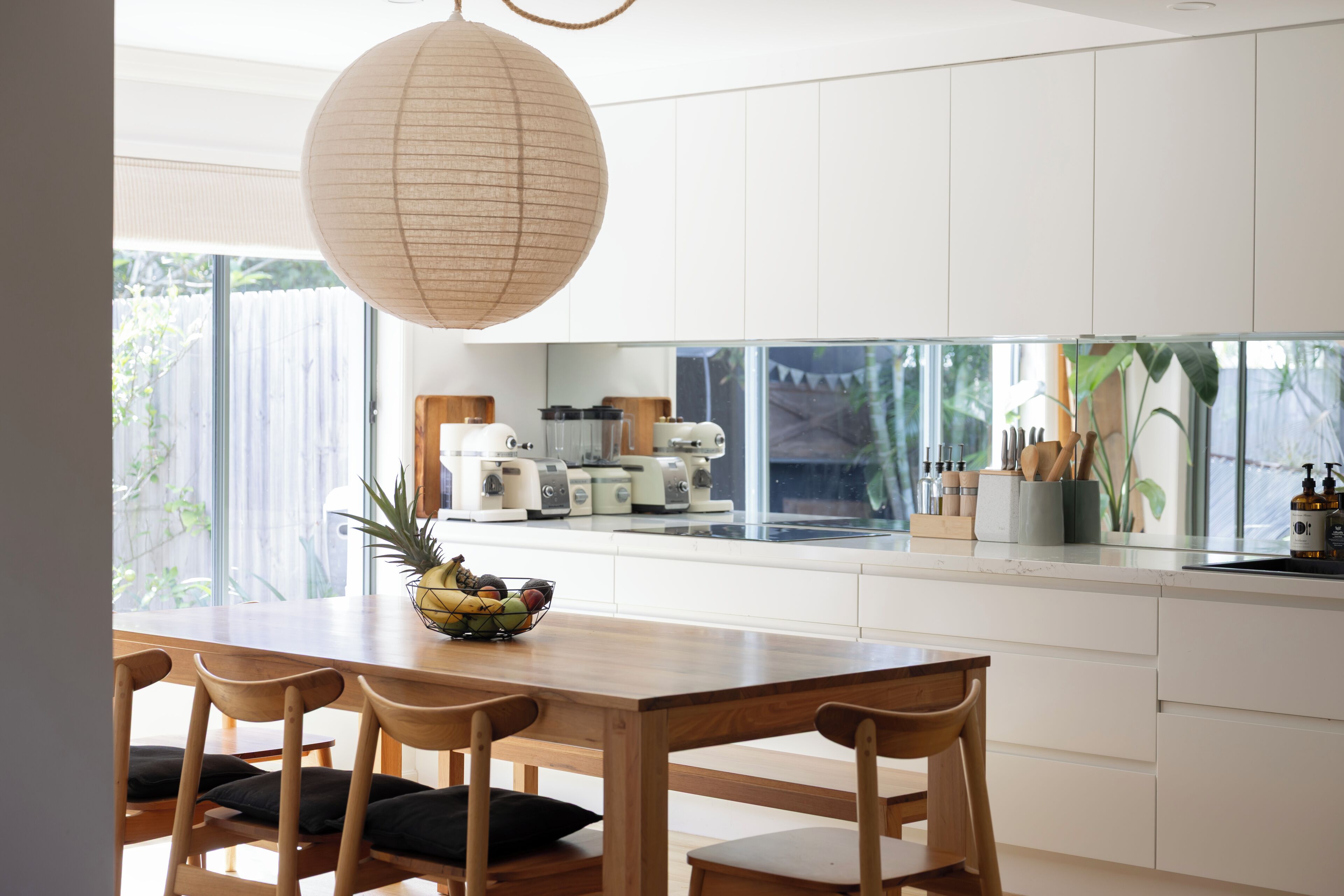A modern kitchen with white cabinets, a wooden dining table, and a large window overlooking a garden.