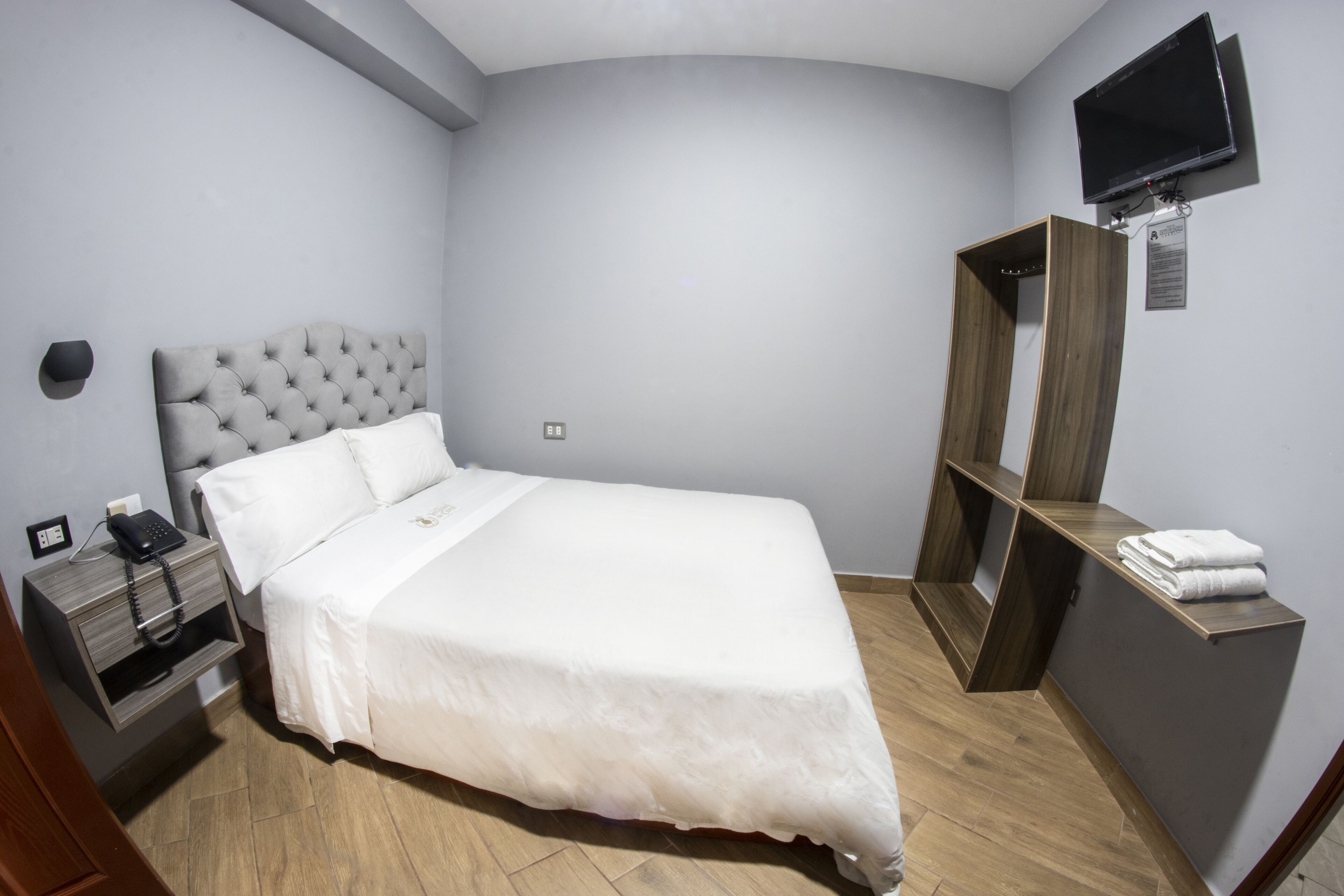 Een keurig opgemaakt slaapkamer met een groot bed, een nachtkastje met een telefoon, een houten nachtkastje, een aan de muur bevestigde televisie en een houten kast met handdoeken.