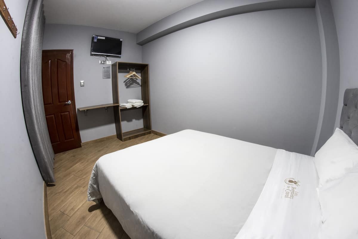 A hotel room with a bed, a television mounted on the wall, a wooden door, and a wardrobe with hanging clothes.